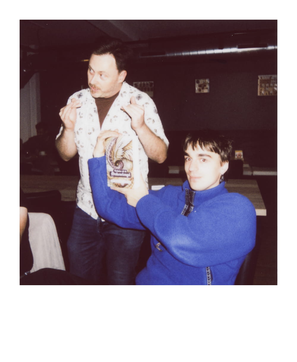 Player holds up a tabletop gaming book while another participant explains something beside the table.